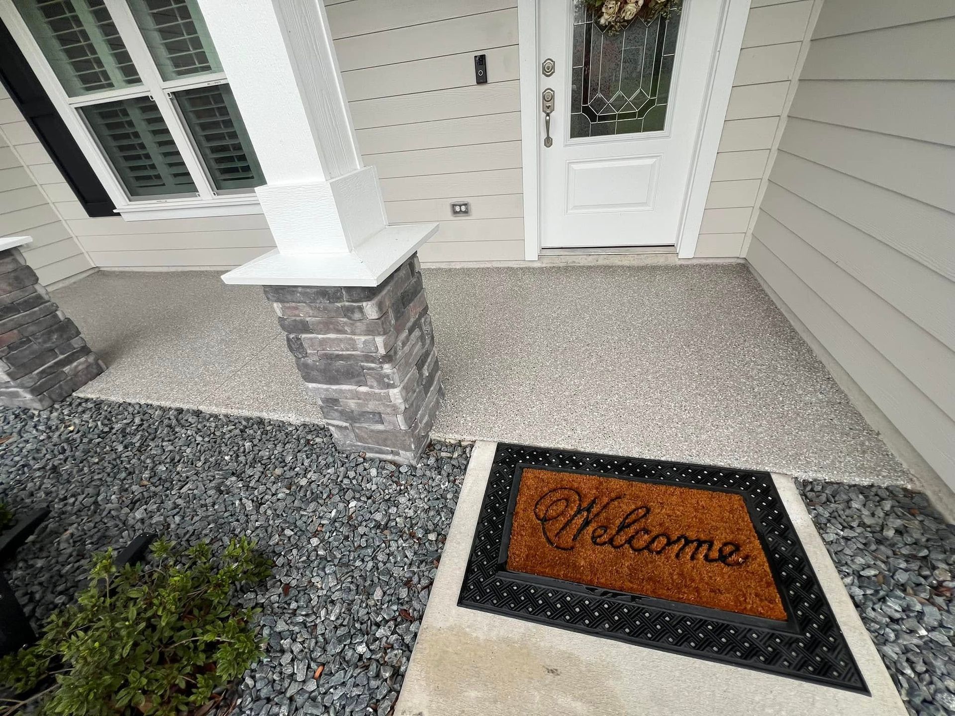 Front porch with welcome mat, stone columns, and gravel landscaping.