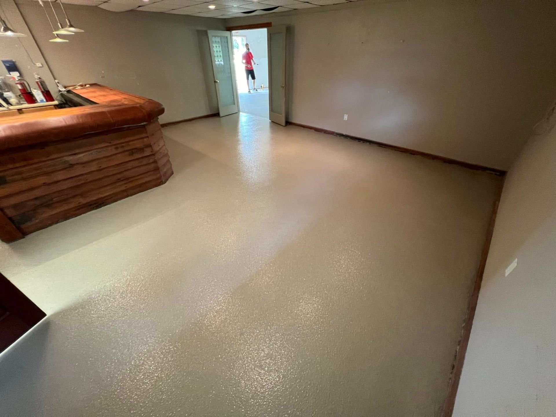 Room with tan speckled floor, wooden bar, and open doorway with person.