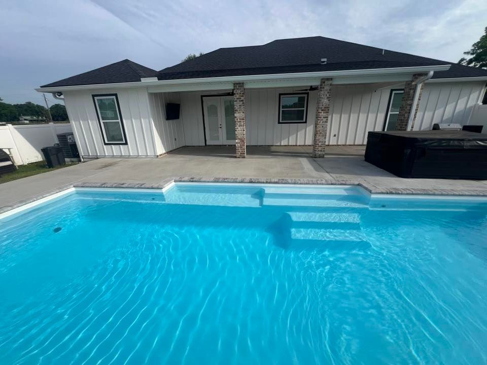 A backyard with a pool, patio, and white house with black roof. Bright blue pool water.