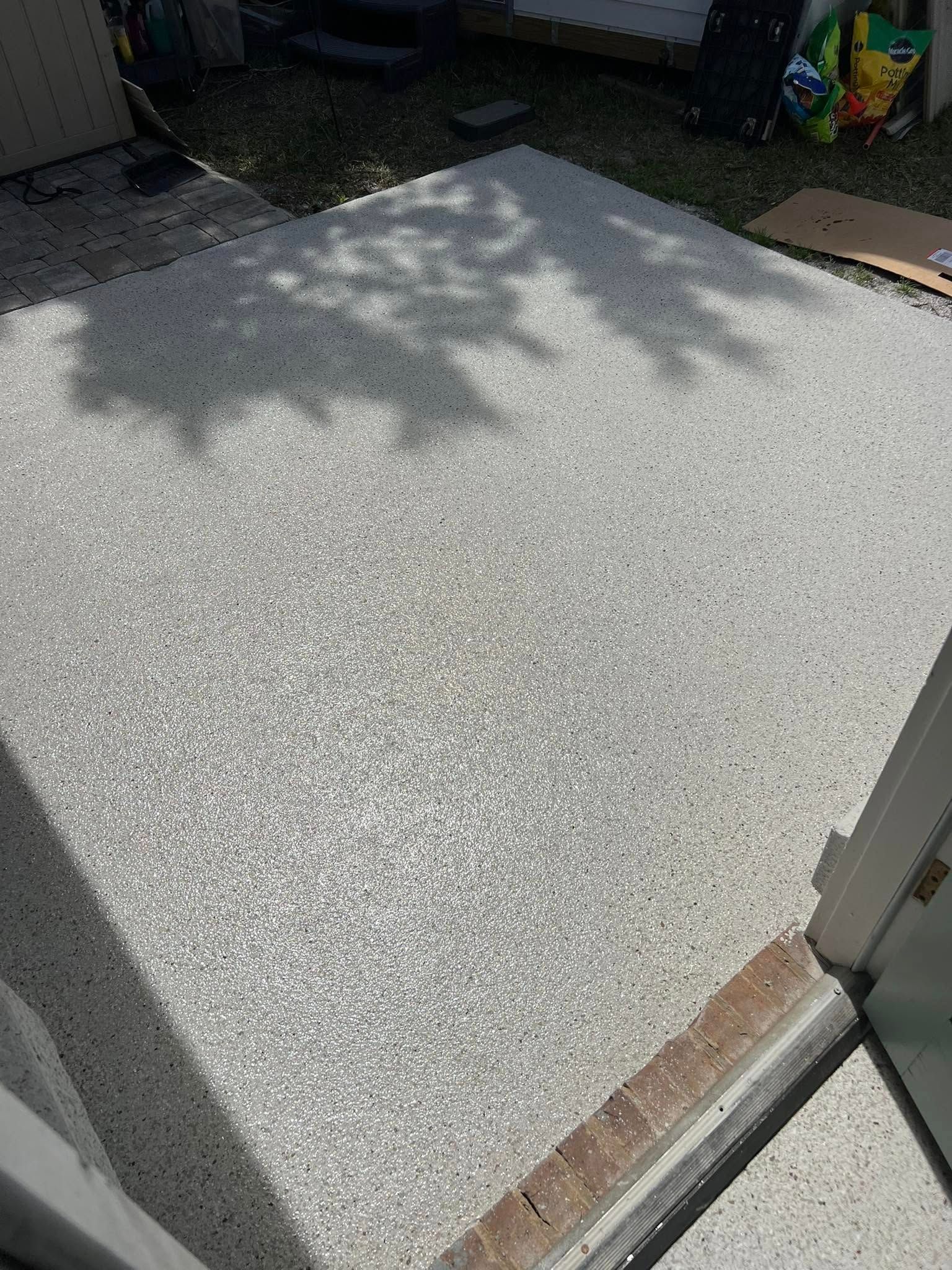 A light gray, speckled concrete patio with shadows from overhead trees next to a sliding glass door.