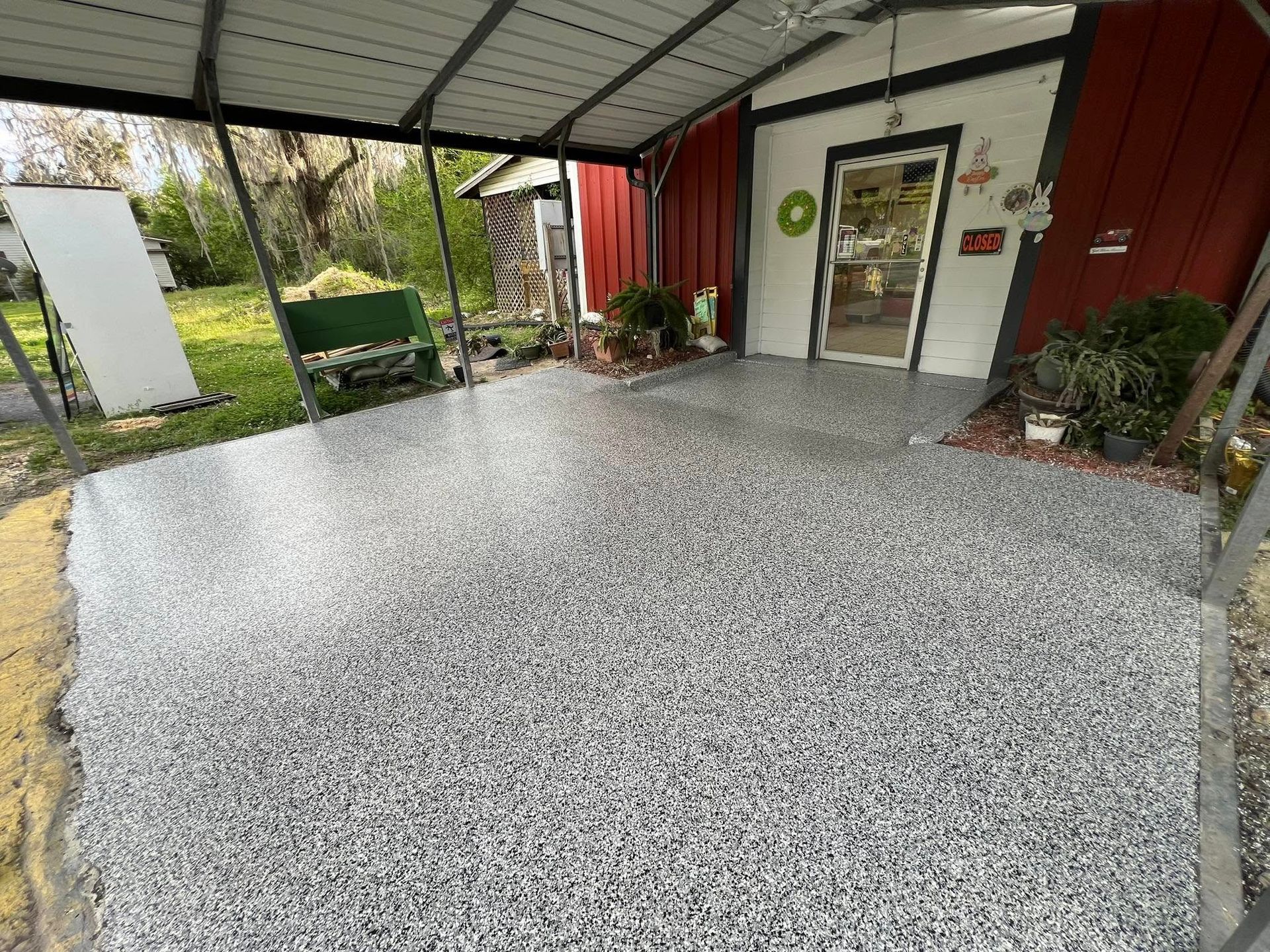 A concrete patio with a speckled gray finish under a metal awning, next to a red building with a glass door.