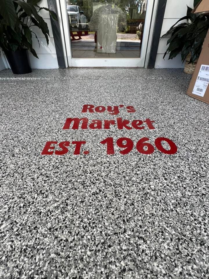 Entrance of Roy's Market. Red logo on gray speckled floor. Doorway reflection. Plants on either side.