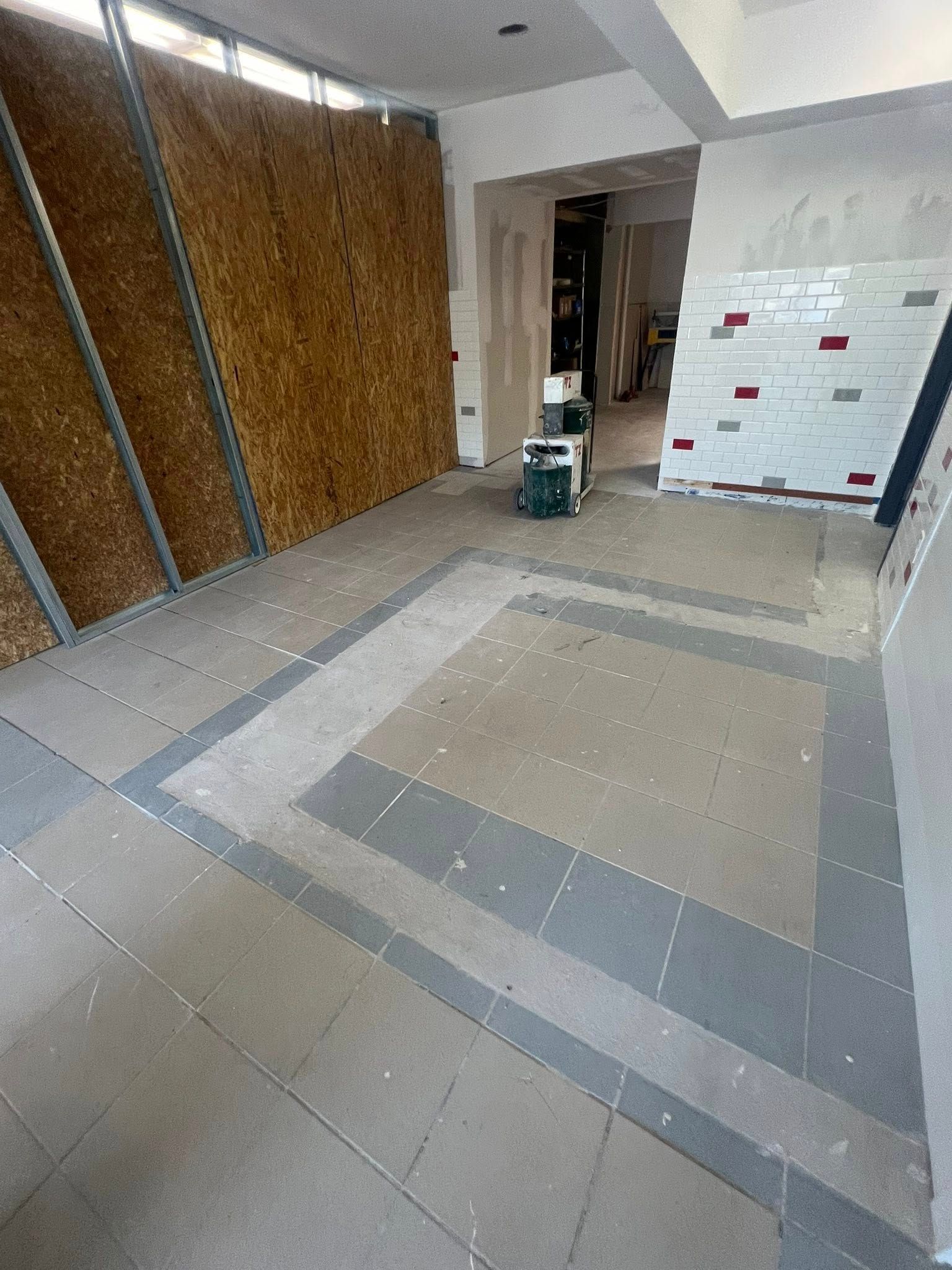 Construction site. Partially tiled floor with exposed framing, wooden wall, and a machine in the center.