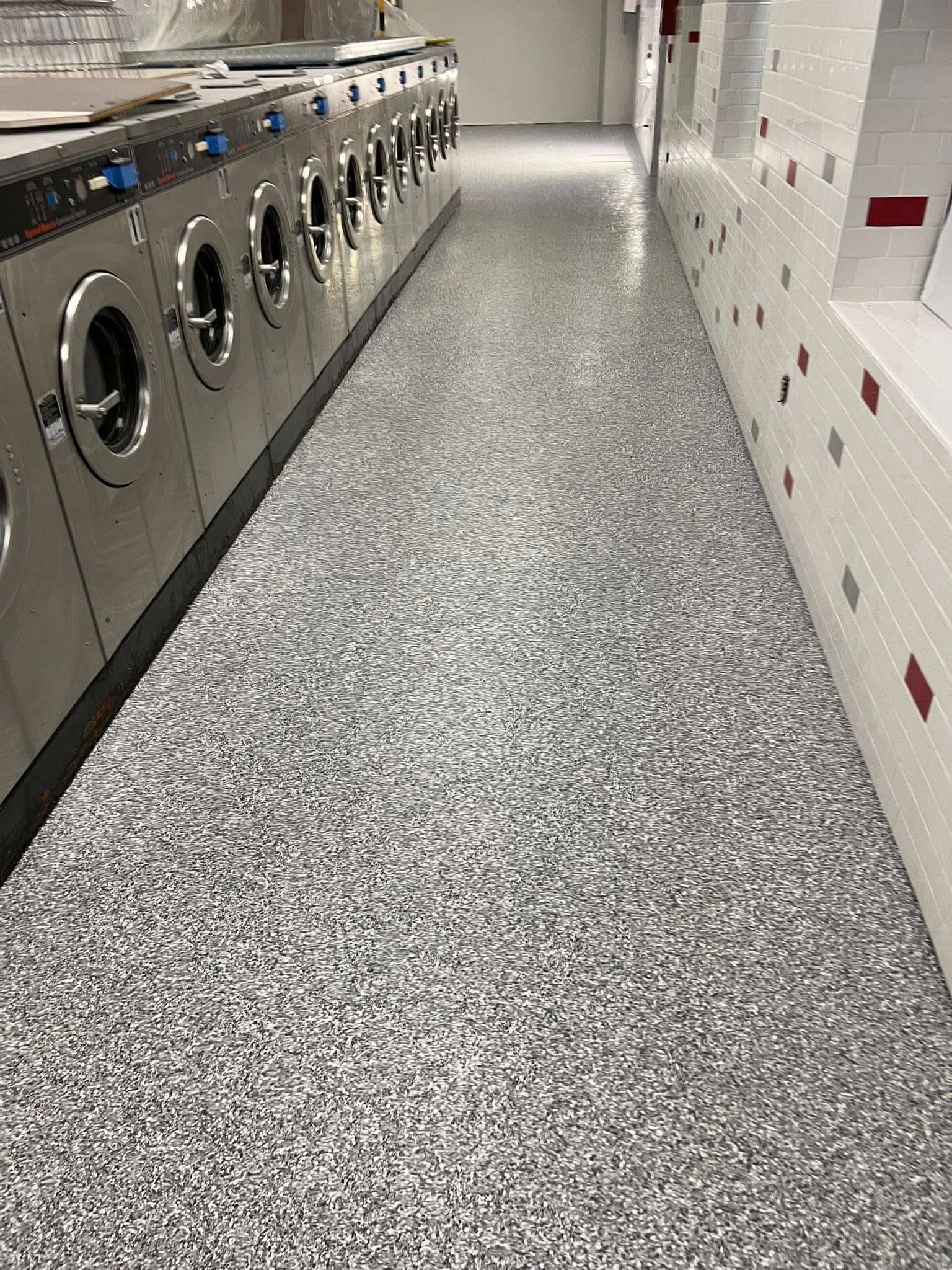 Row of washing machines in a laundromat, gray and white speckled flooring, white tiled wall with red accents.