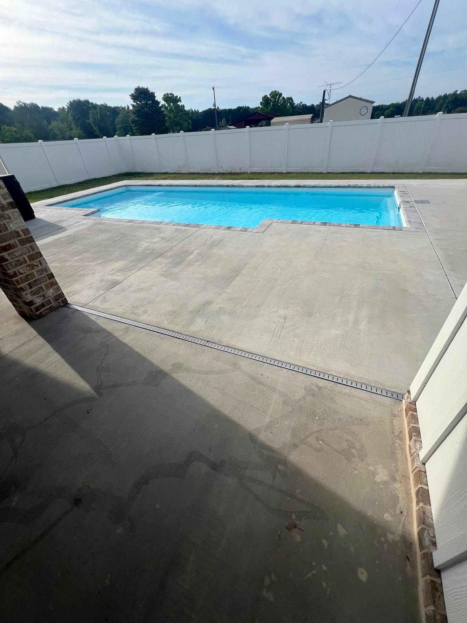 A rectangular swimming pool surrounded by concrete patio, white fence, and blue sky.