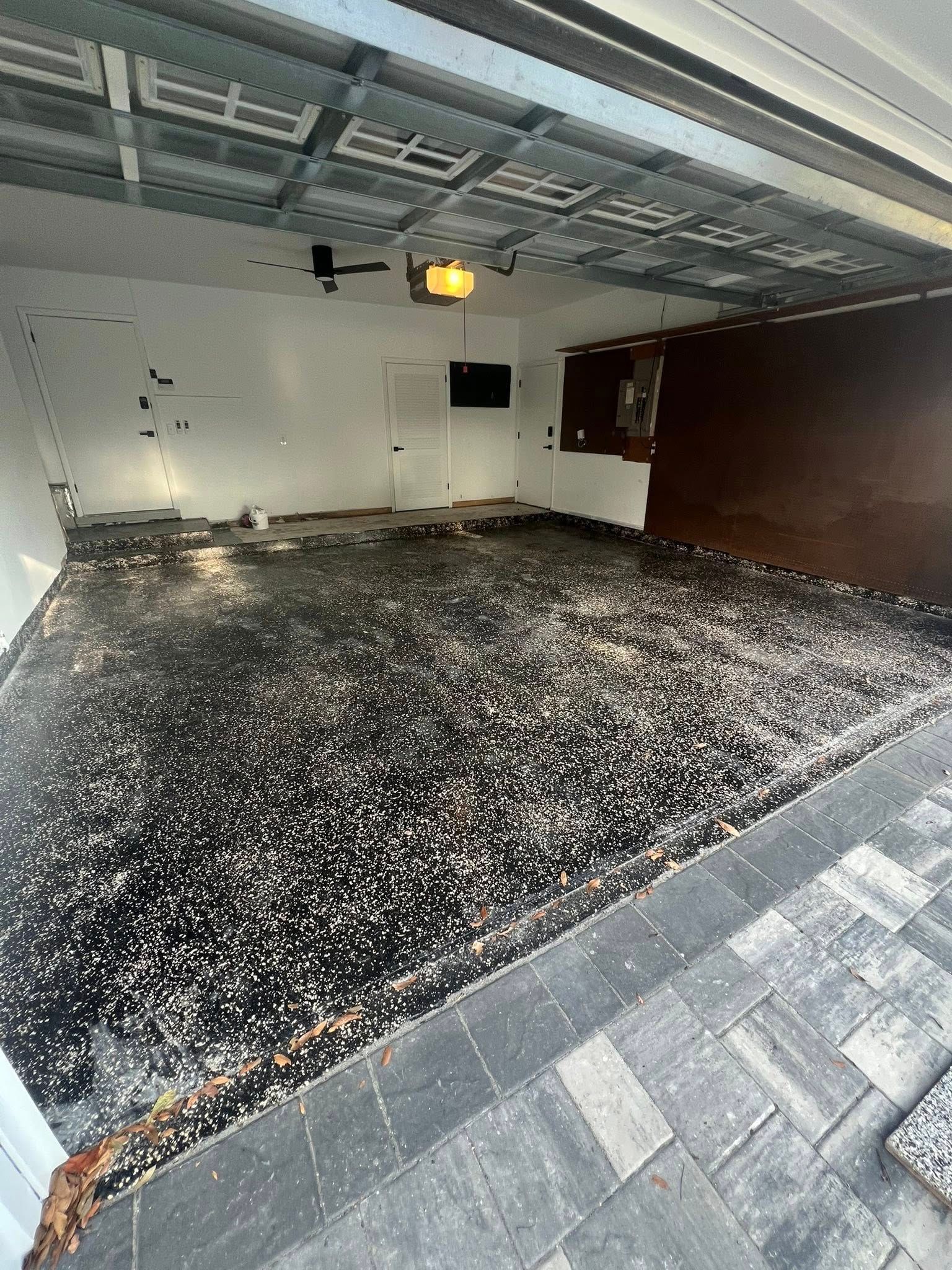 Garage interior with damaged floor covered in dark debris. White walls, open door frame, overhead lighting.