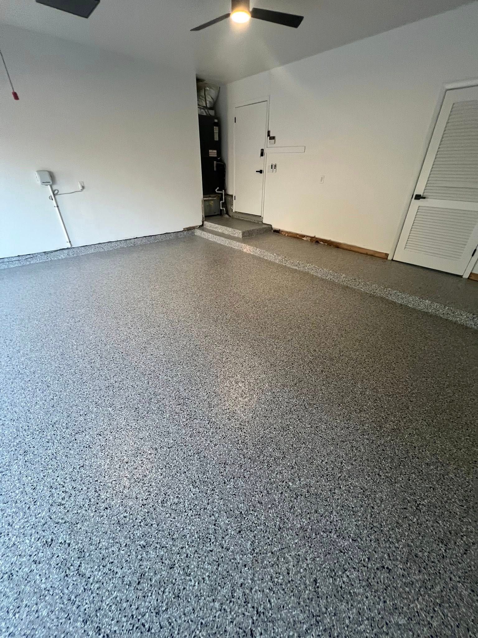 Empty garage with speckled gray floor and white walls. A staircase leads to a dark doorway in the back.