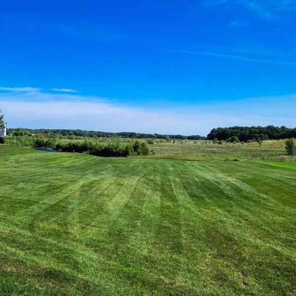 Lush green lawn with mowing stripes stretches toward a field and treeline under a bright blue sky.