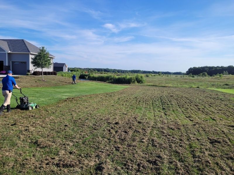 Two people mowing a large lawn with a house in the background on a sunny day.