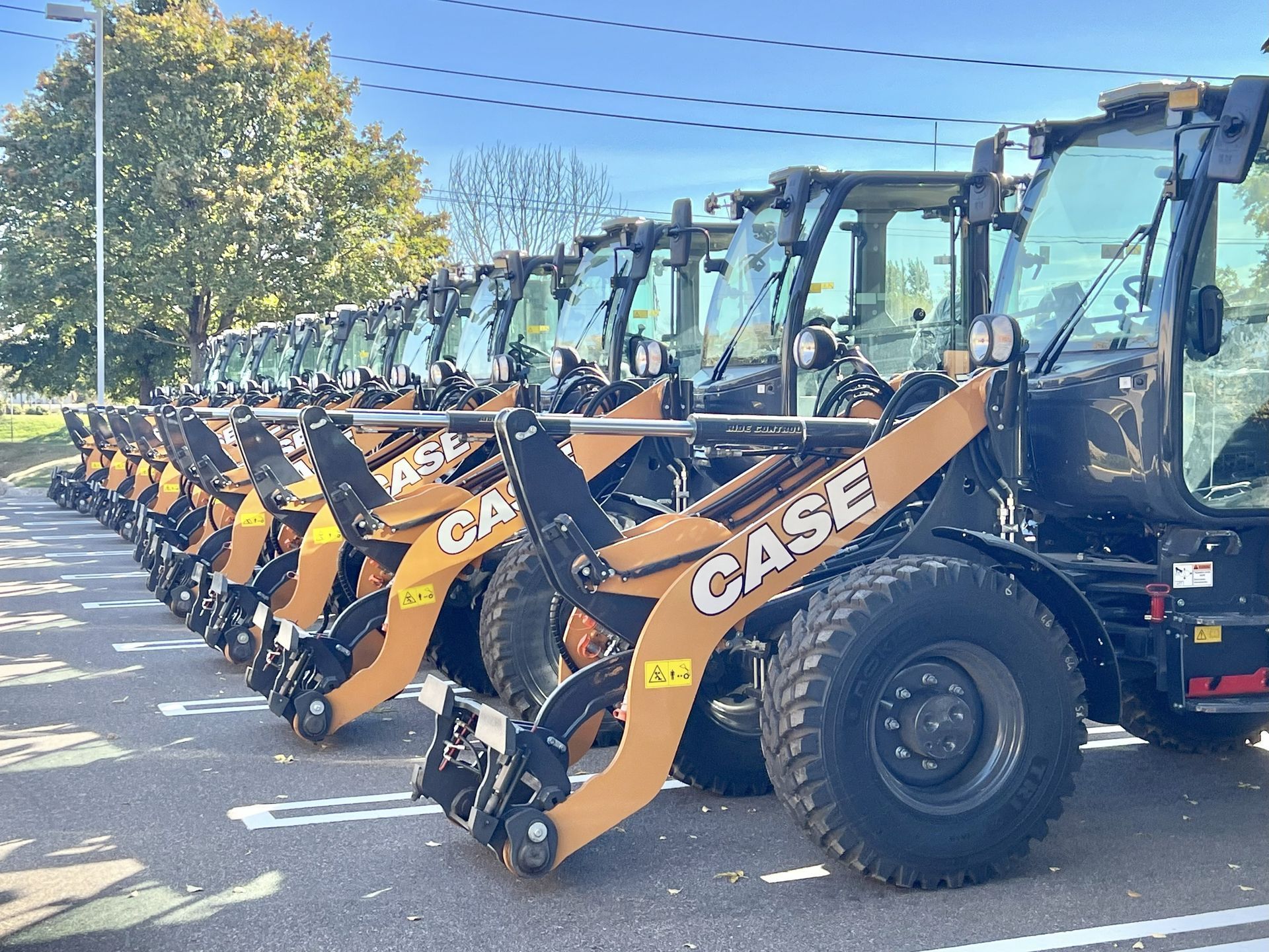 Row of new Case skid steer loaders, parked in a lot, with raised orange loader arms, under a bright sky.