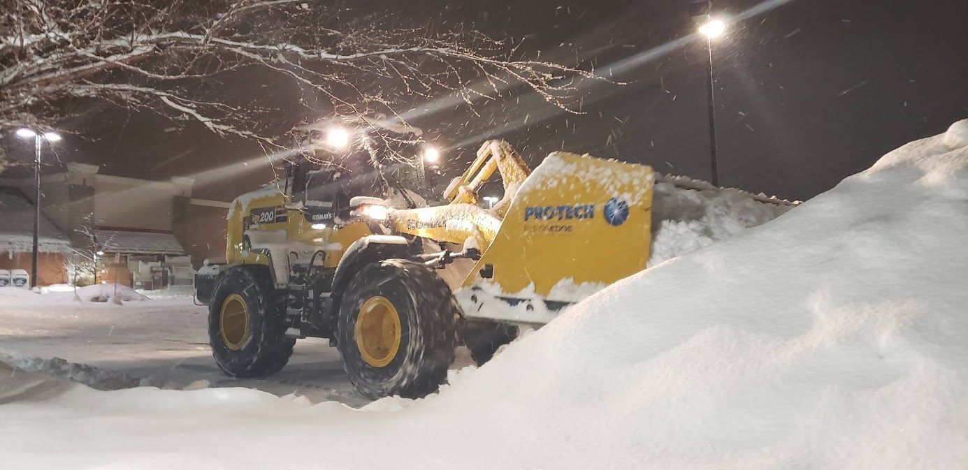 A yellow snowplow clearing snow at night. The vehicle has large wheels and is next to a large pile of snow.