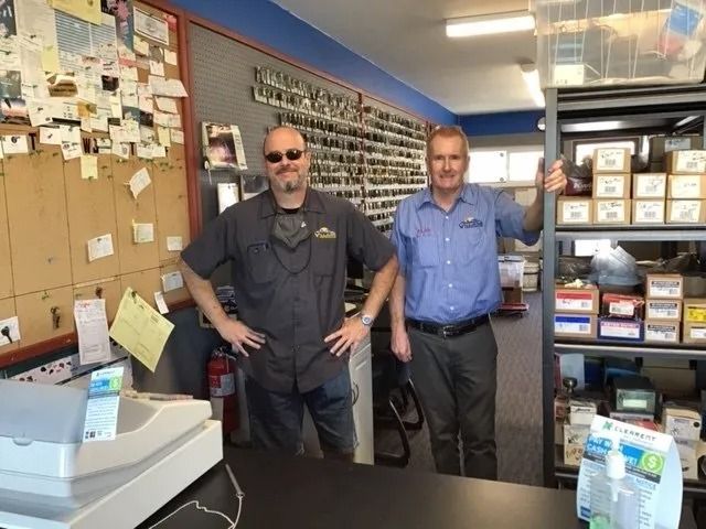 Two men behind a shop counter. One in sunglasses, one giving a thumbs-up. Keys on the wall, shelves with boxes.