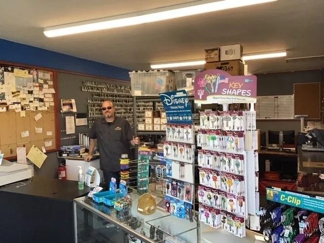 Man in a locksmith shop stands behind a counter. Keys on display, boxes, and fluorescent lights are visible.