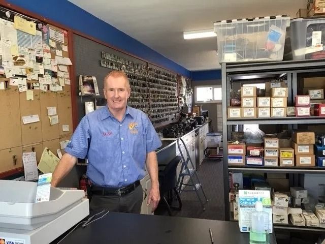 Man in blue shirt stands at a counter in a hardware store. Shelves of boxes and parts in background.