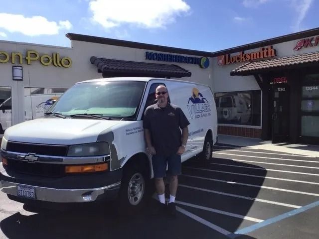 Man standing next to a white service van in front of a shopping center with storefronts. Sunny day.