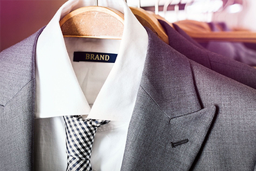 Gray suit jacket with white shirt and patterned tie on a hanger, with a black “BRAND” label