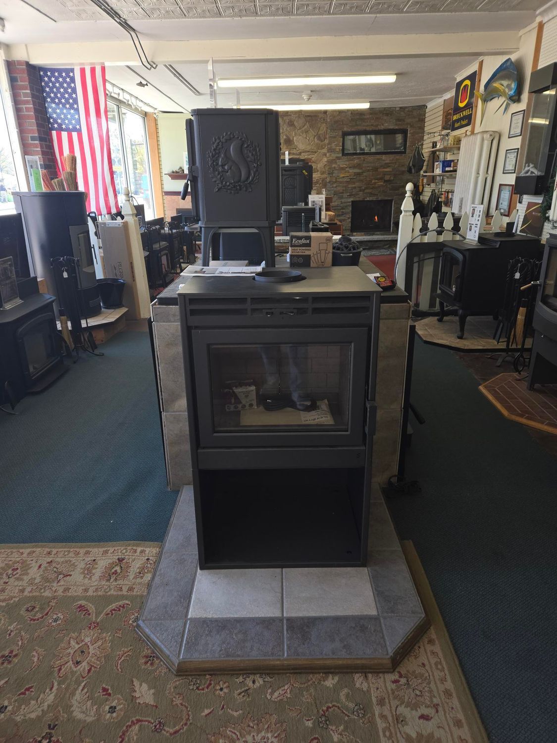 Wood-burning stove centered in a showroom, surrounded by rugs, furniture, and American flags.