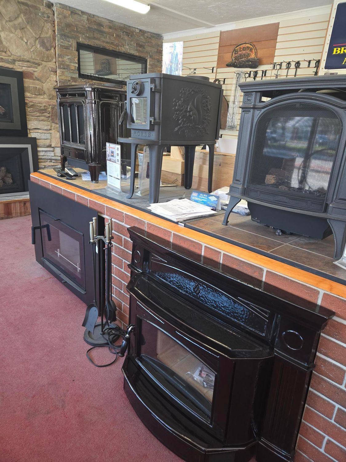A brick hearth display with several black wood stoves and fireplace inserts in a showroom