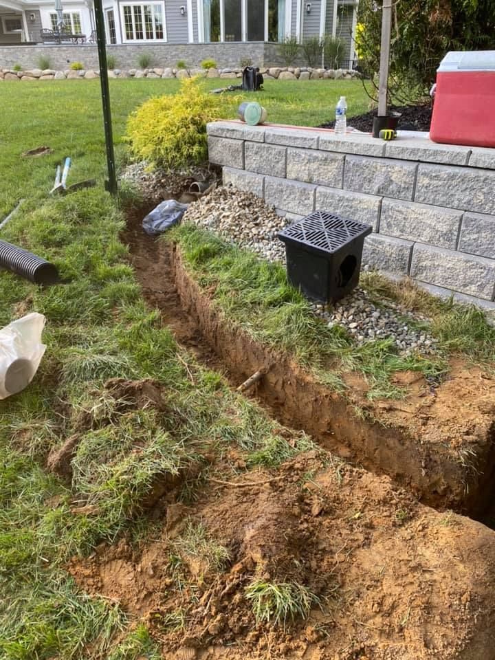 A trench is being dug in the grass next to a brick wall.