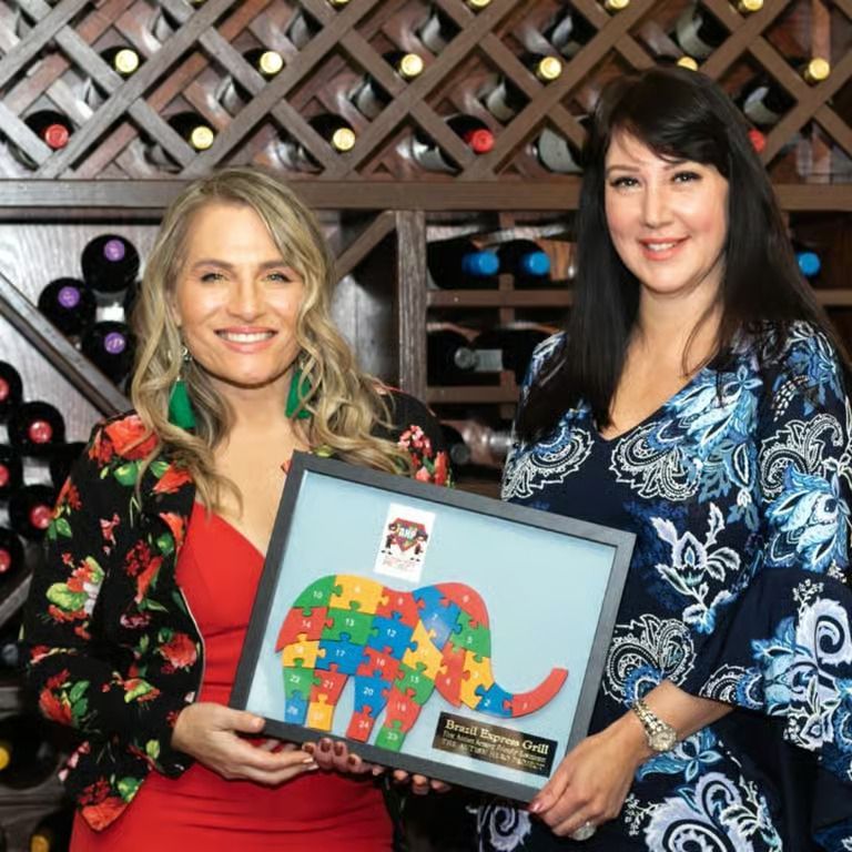 Two women holding a framed colorful elephant artwork in front of a wine rack