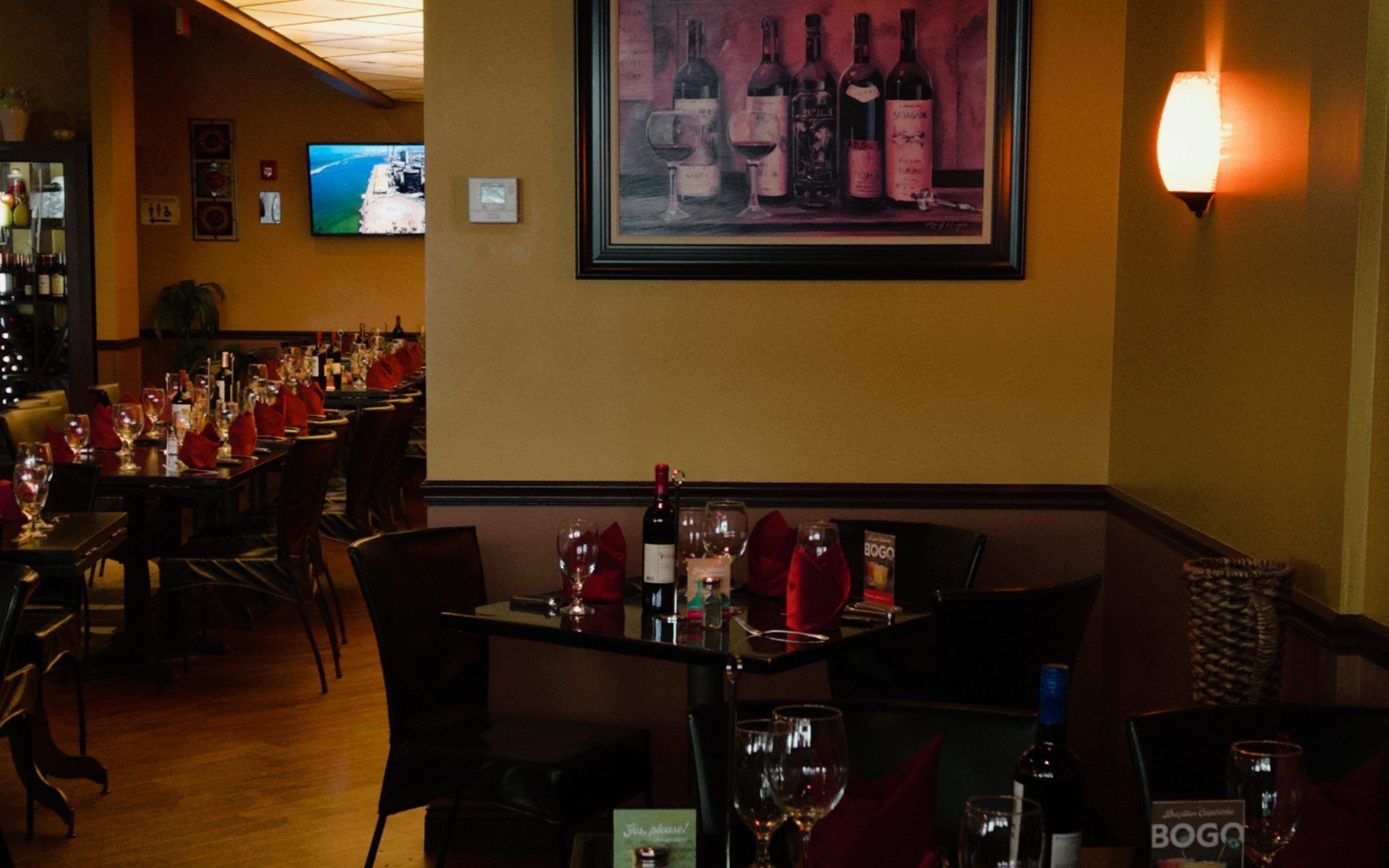 Dimly lit restaurant dining area with set tables, red accents, and wall art near a TV.