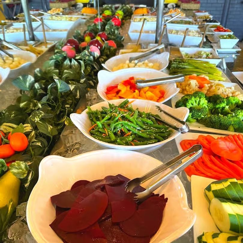 Buffet table with assorted salads, vegetables, and serving tongs in white bowls
