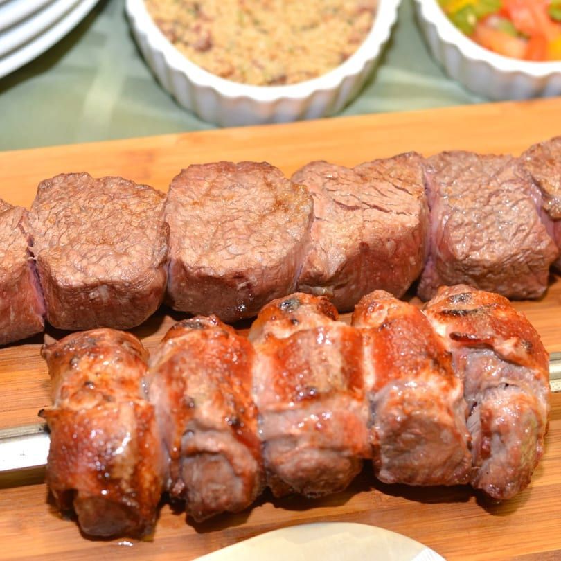 Grilled skewered beef cubes on a wooden board with side dishes in the background