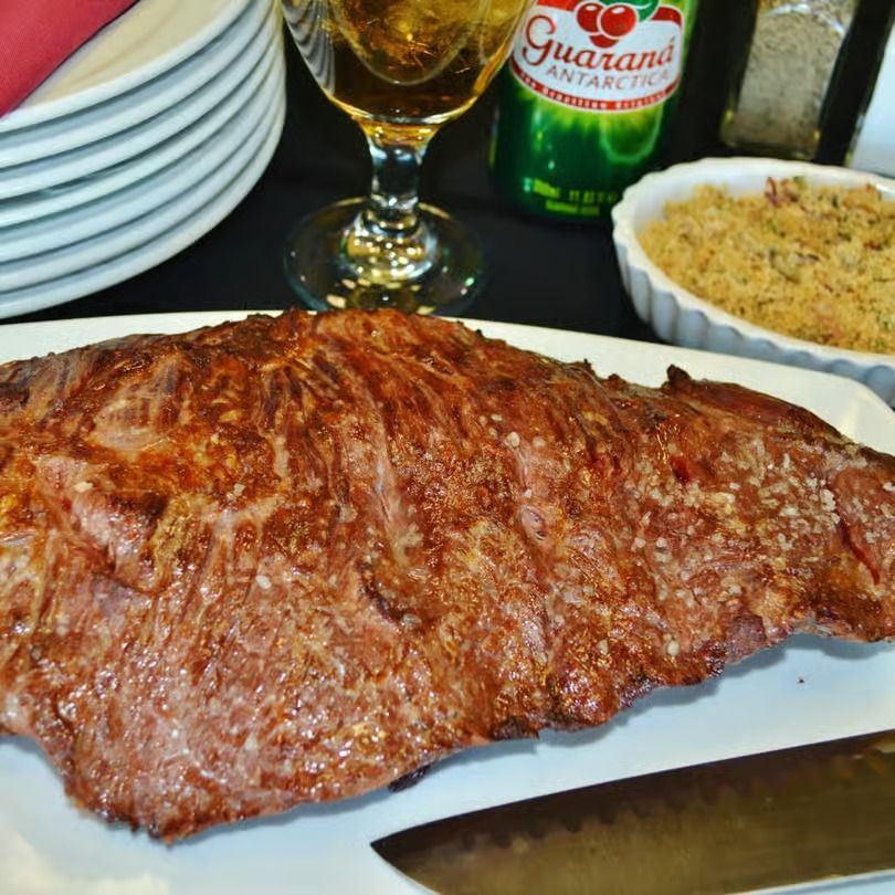Grilled steak on a white plate with rice, Guaraná soda, and stacked plates in the background