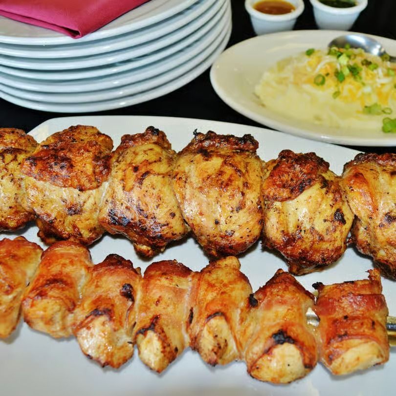 Grilled chicken skewers on a white platter with pasta and stacked plates in the background