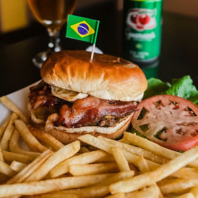 Burger and fries with a Brazilian flag toothpick, tomato slice, and soda can in the background