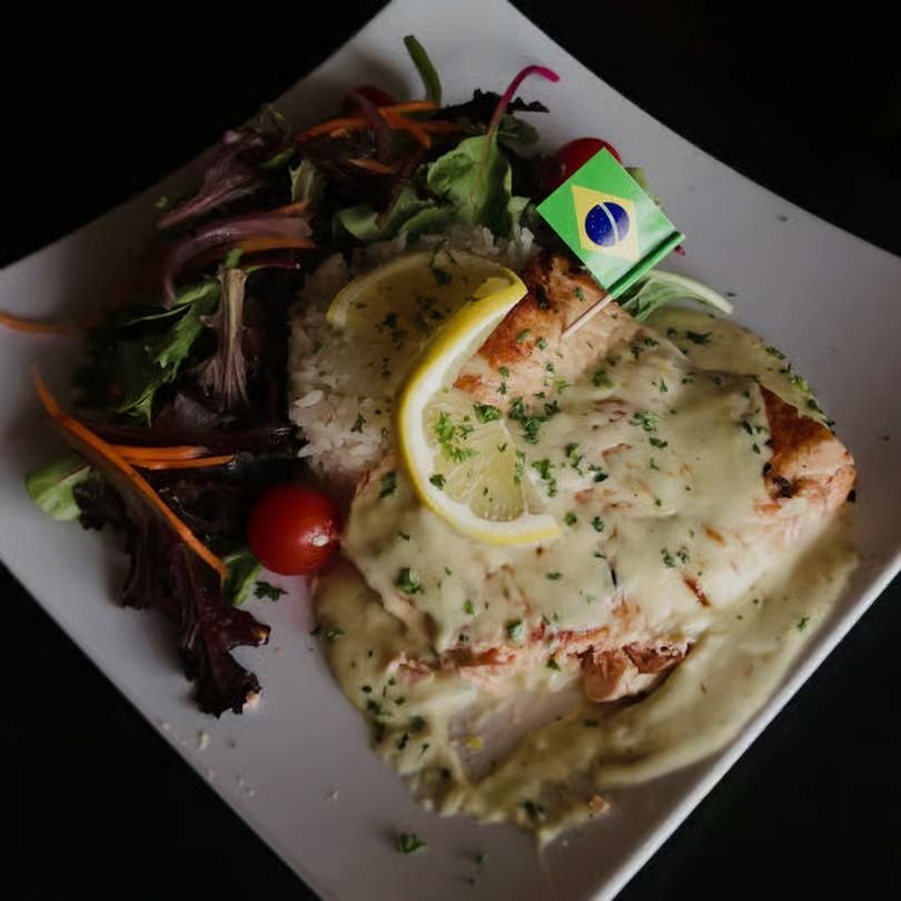 Creamy grilled fish with lemon on a white plate, served with mixed greens and cherry tomato, topped with a tiny flag.