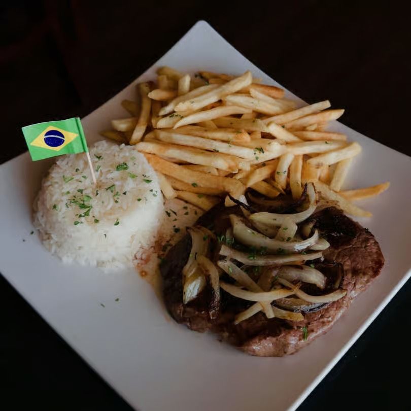 Brazilian plate with rice, fries, and grilled steak topped with onions and a small Brazil flag
