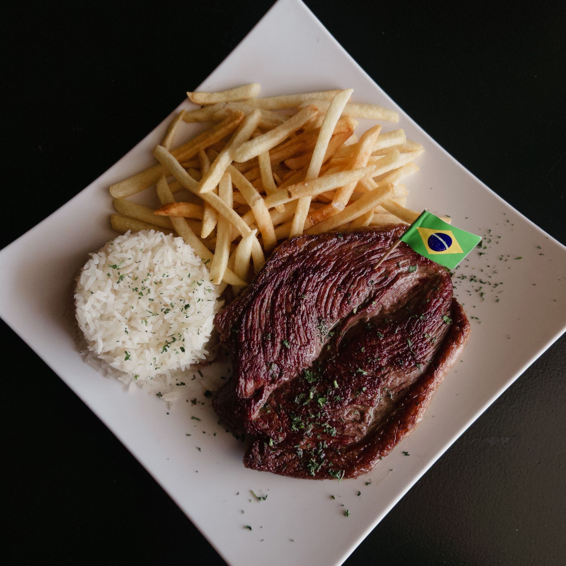 Grilled steak with rice, fries, and a small Brazilian flag on a square white plate.