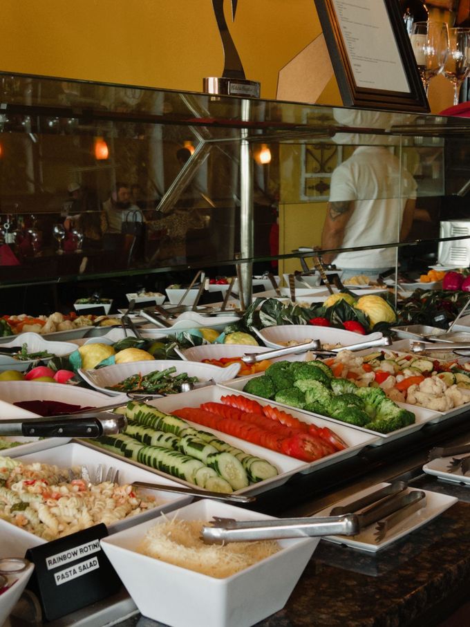 Self-serve salad bar with trays of vegetables, cheeses, and toppings under glass display
