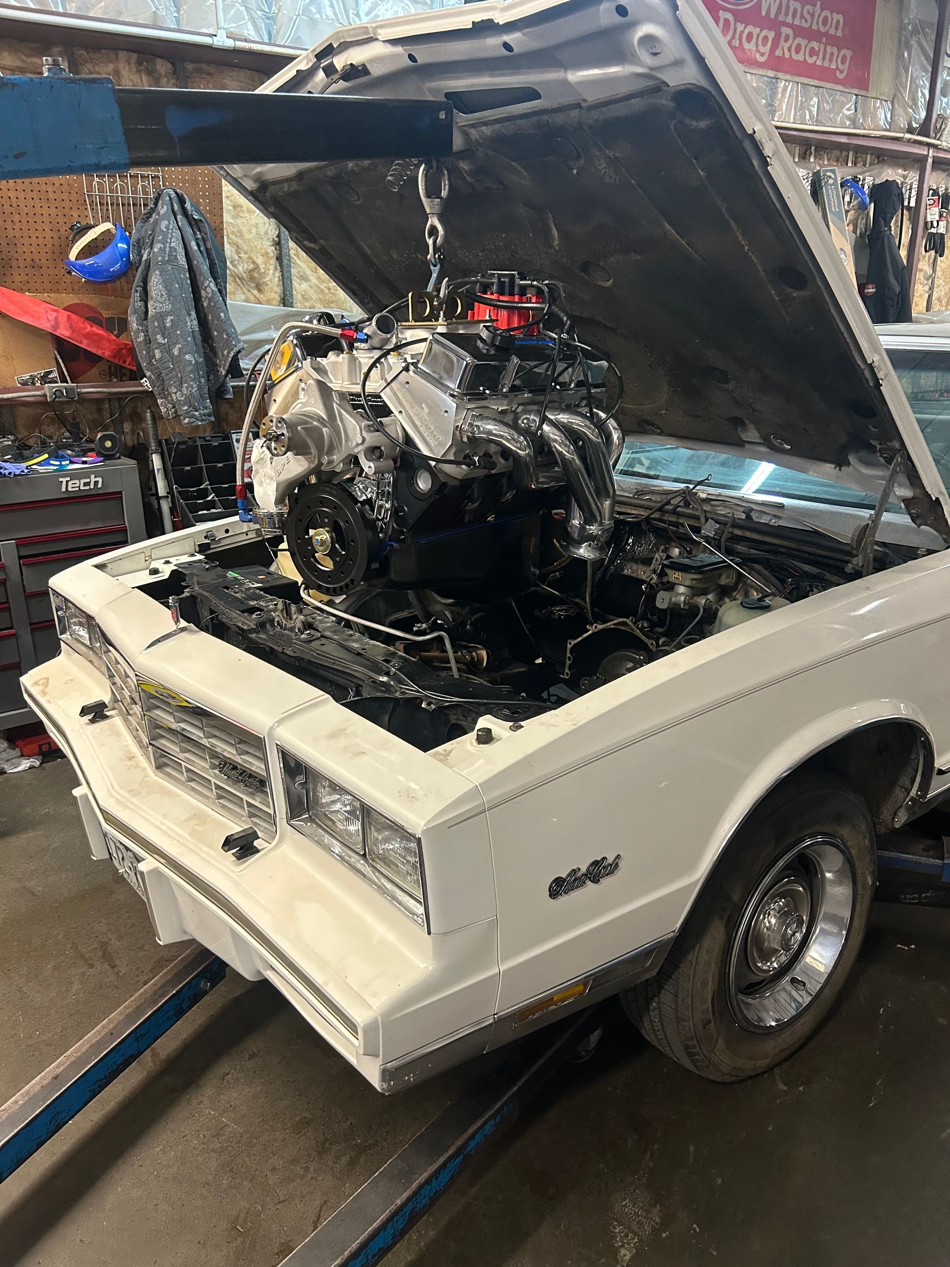A white car with open hood, engine being lowered by a hoist in a garage.