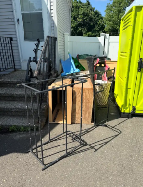 Metal shelving unit and a table of miscellaneous items on a driveway beside a house and bright green portable toilet.