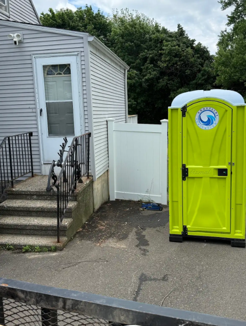 Bright yellow portable toilet beside a white house and fence, with stairs and parked bicycles nearby