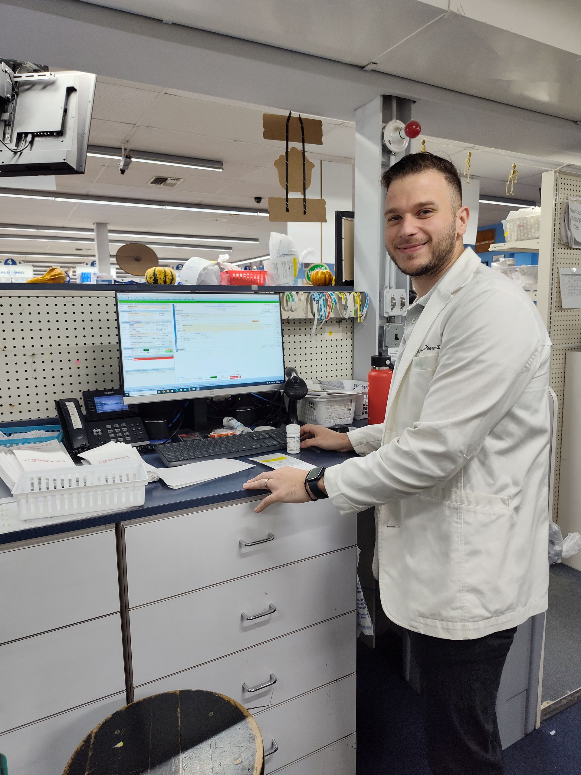 A man in a lab coat is standing in front of a computer in a pharmacy.