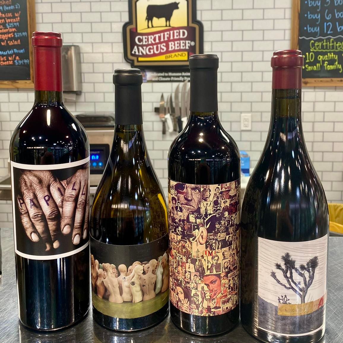 Four wine bottles with unique labels, set on a countertop in a store, near a Certified Angus Beef sign.