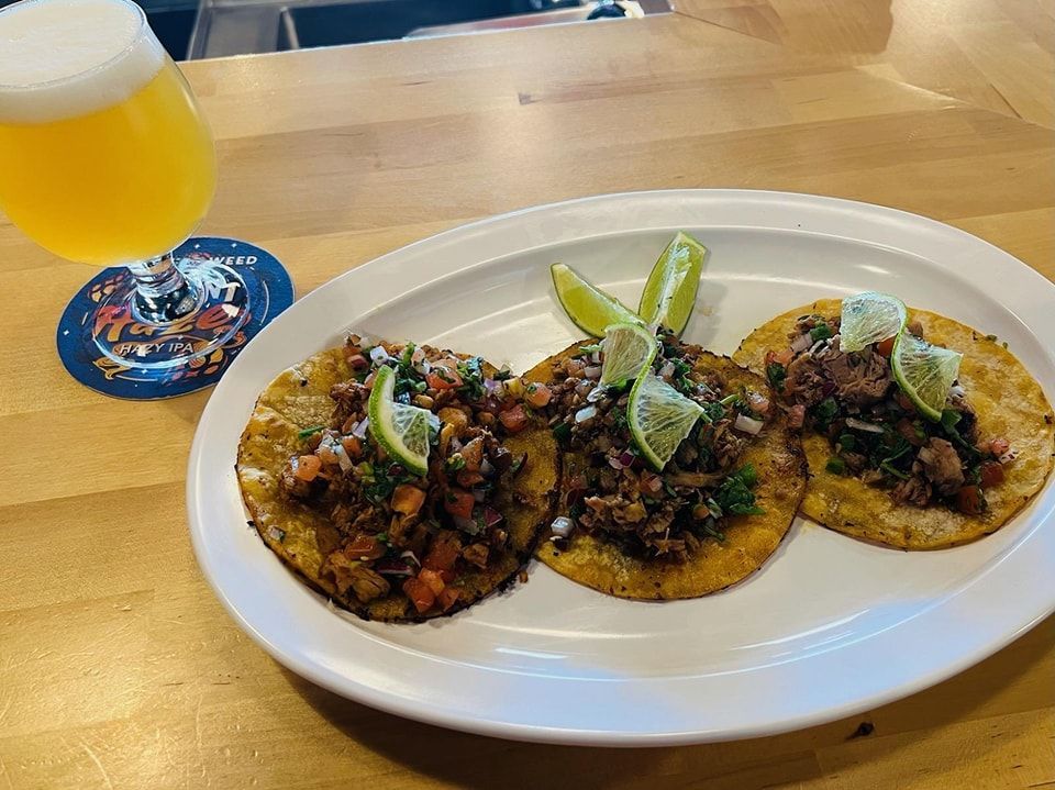 Three tacos with lime wedges on a white plate, beside a glass of beer on a coaster.