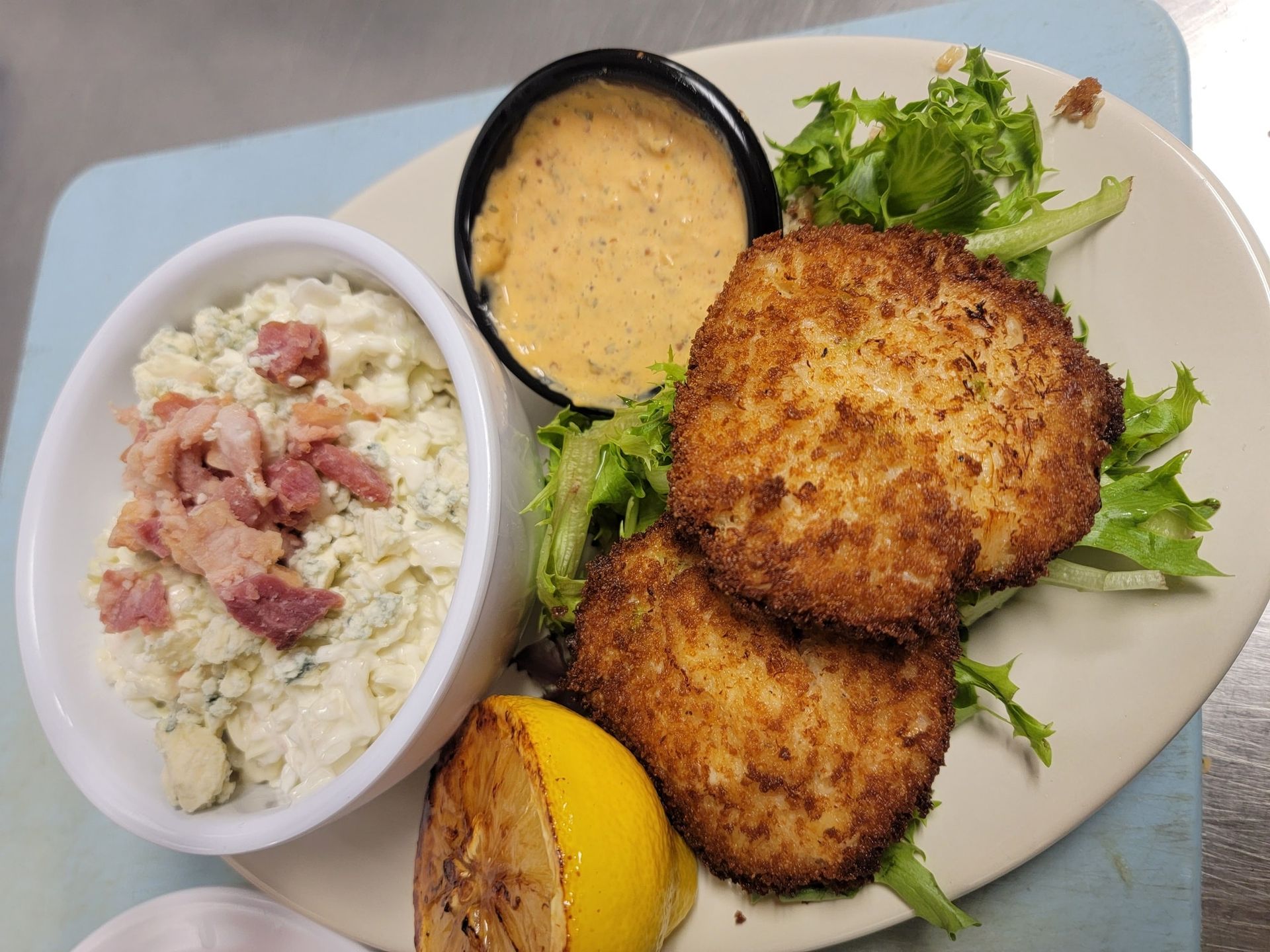 Two crab cakes, salad, lemon wedge, dip, and blue cheese/bacon salad on a plate.