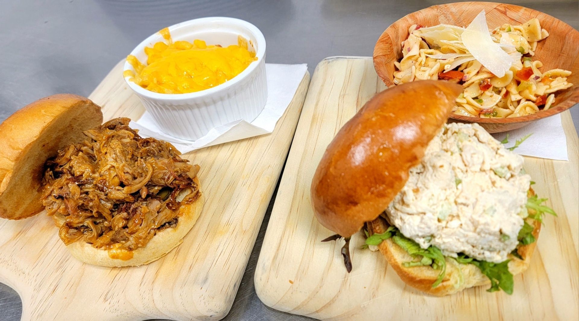 Two sandwiches with sides on wooden boards: pulled pork and chicken salad.