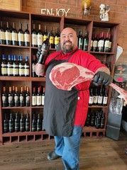 Man holding a large steak and a bottle of wine in a wine shop.