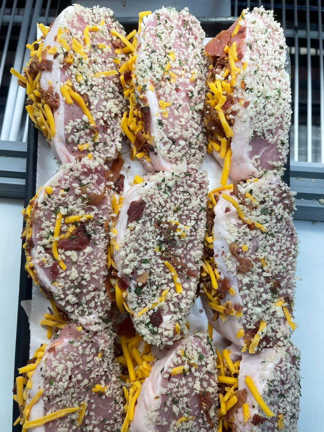 Raw pork chops coated in breadcrumbs, shredded cheese, and bacon on a baking sheet.