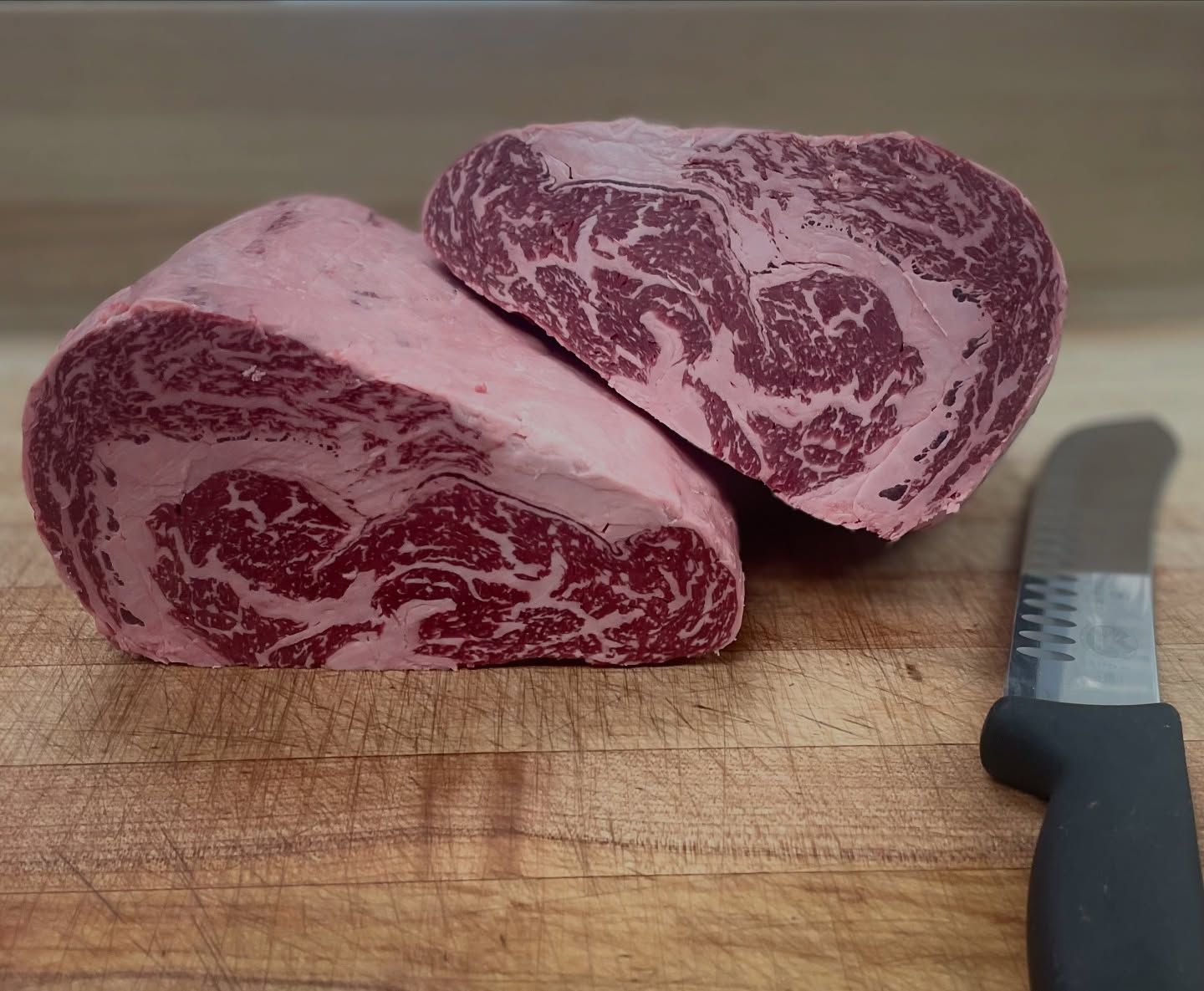 Two raw steaks with heavy marbling sit on a wooden surface, near a knife.