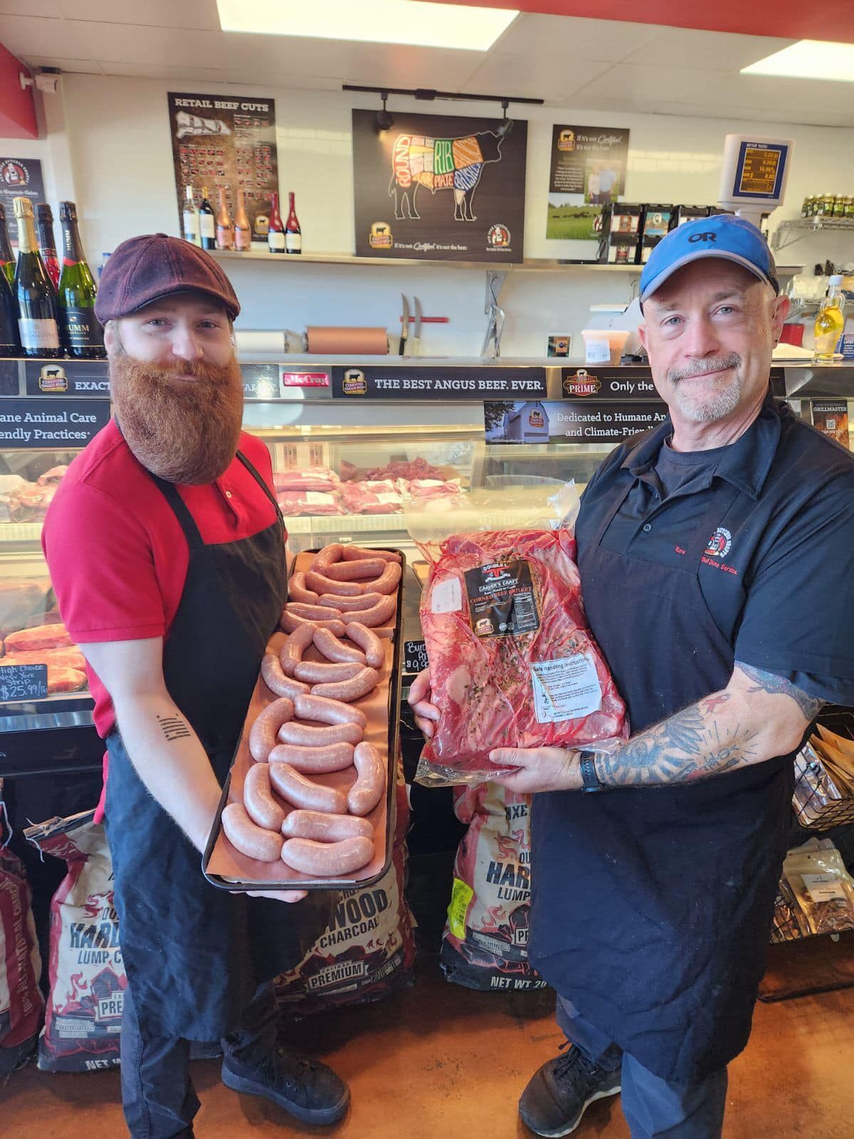 Two men in butcher shop holding tray of sausages and a large cut of meat.