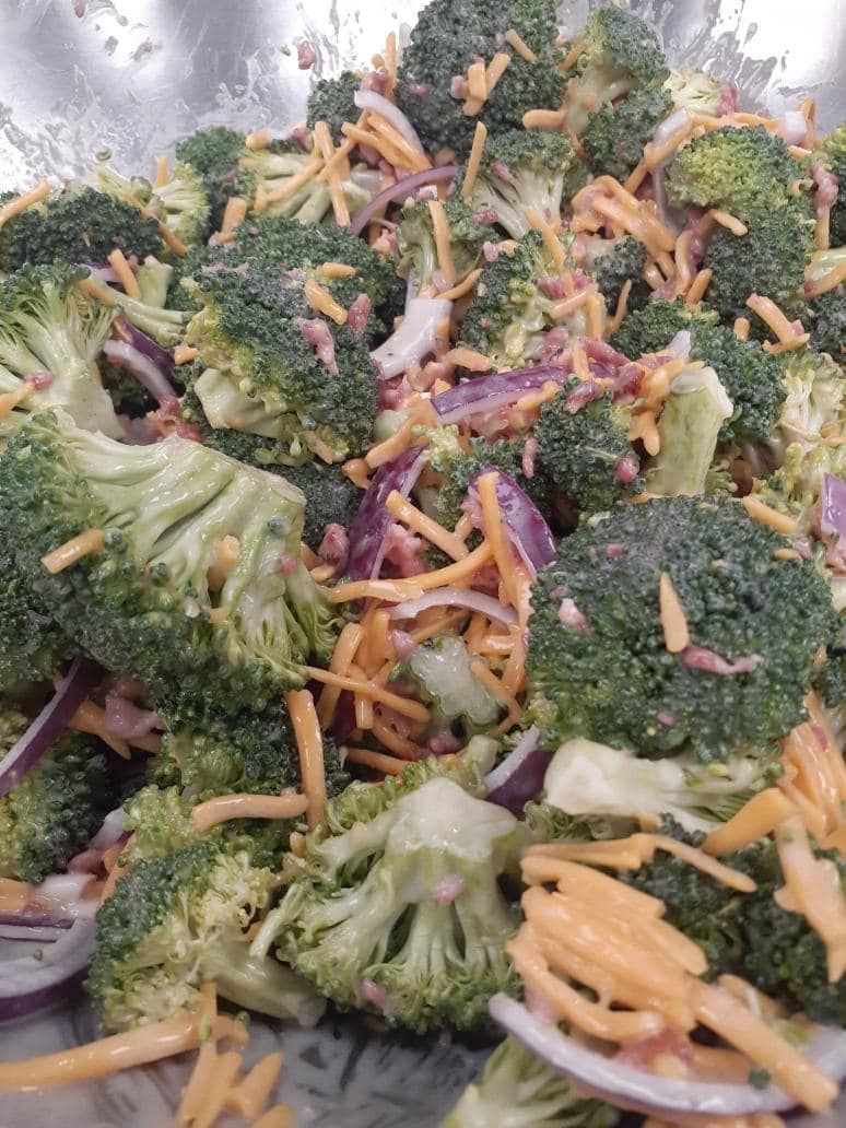 Broccoli salad: green broccoli florets, red onion, shredded cheddar cheese, in a metal bowl.