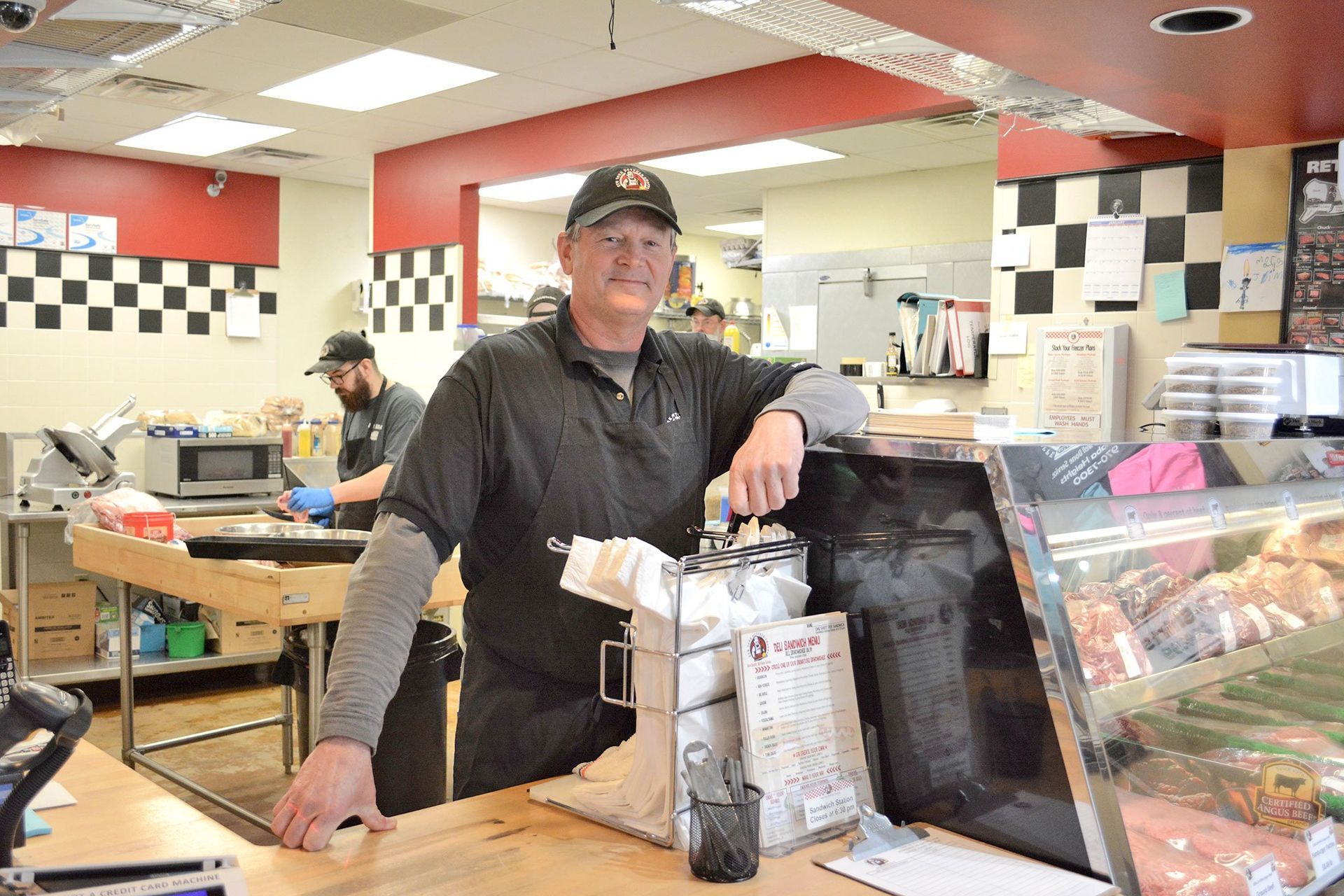 Man in butcher shop, leaning on counter, smiling. Employee in background preparing food.