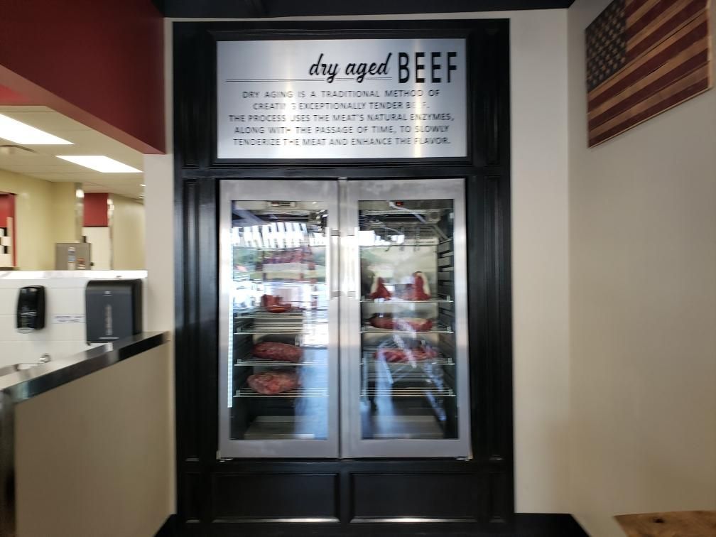 Dry-aged beef display case inside a restaurant, with glass doors, text above, and an American flag nearby.