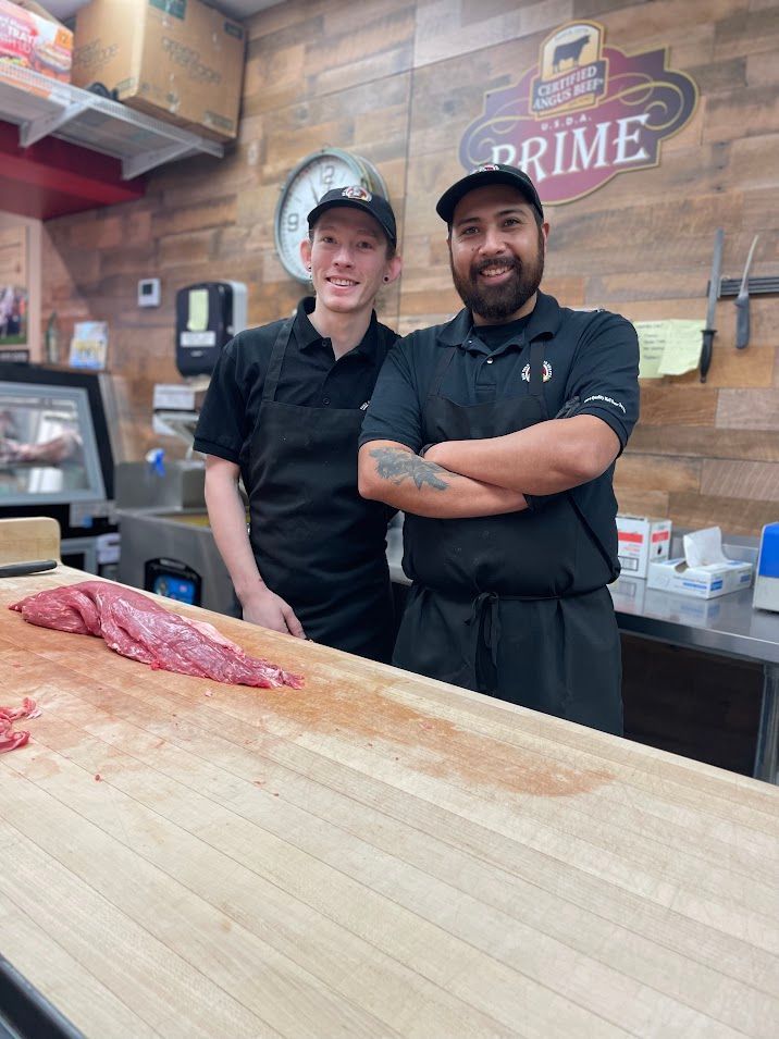 Two men in butcher shop, one smiling, one with crossed arms. A meat counter and product sign.